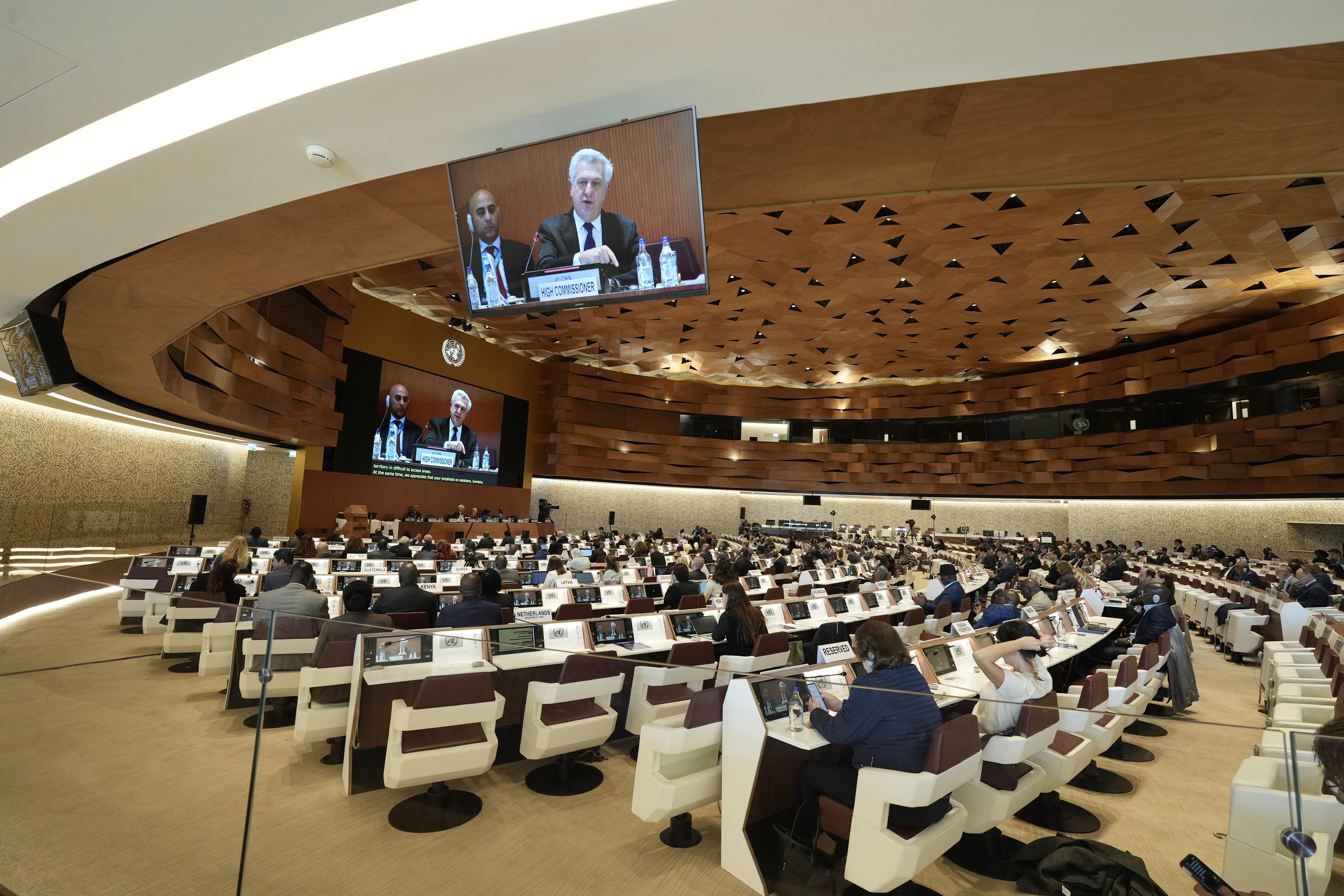 A modern conference room filled with attendees, facing large screens that display speakers at the front during a formal meeting of the UNHCR Executive Committee.