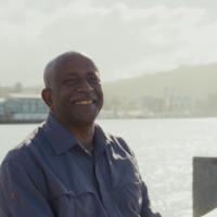 Person standing near a waterfront with a city skyline and hills in the background on a bright day.