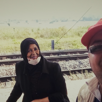 Two people standing on a train platform near railway tracks, with grassy fields and trees visible in the background.