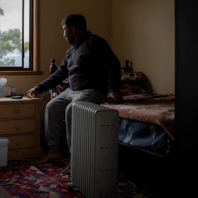 Person sitting on a bed in a small bedroom with a portable heater, nightstand, and window showing trees.