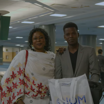 Two people standing in an airport baggage claim area near a sign with the number 6, holding a white bag.