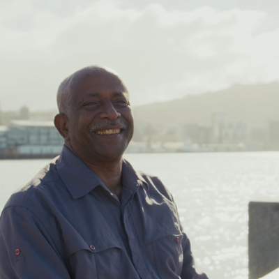 Person standing near a waterfront with a city skyline and hills in the background on a bright day.