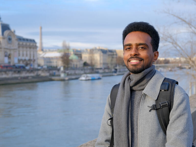Person standing by a river in a city, wearing a grey coat and scarf with a backpack, with historic buildings and a bridge blurred in the background.