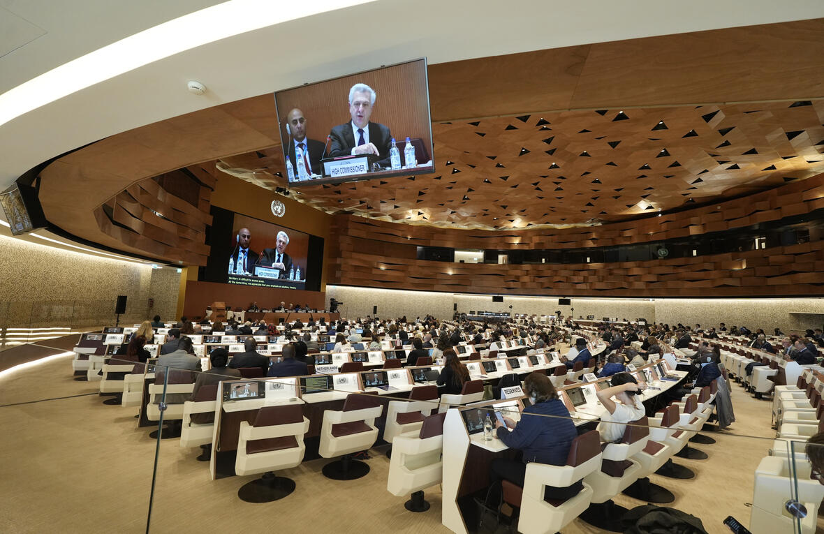 A modern conference room filled with attendees, facing large screens that display speakers at the front during a formal meeting of the UNHCR Executive Committee.