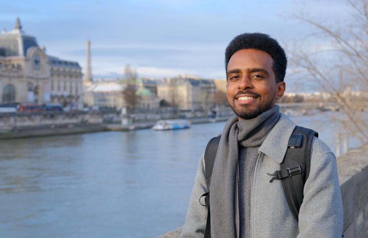 Person standing by a river in a city, wearing a grey coat and scarf with a backpack, with historic buildings and a bridge blurred in the background.